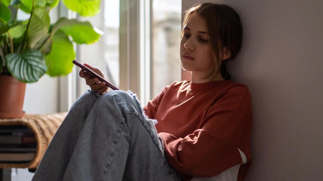 A teenage girl sitting on the floor against a wall, looking at her phone